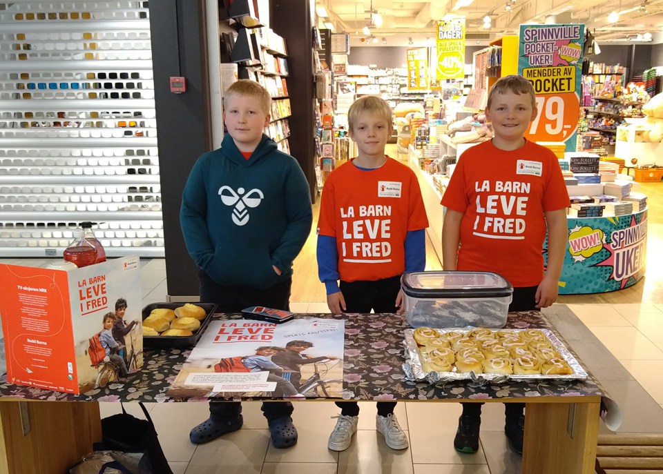 Tre unge gutter står bak et bord med boller og TV-aksjonsplakater. De har på seg tskjorter fra TV-aksjonen.