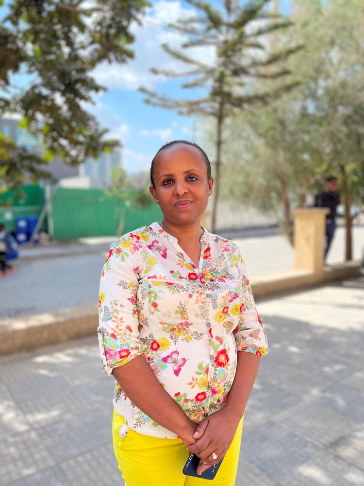 Kvinne står ute med blomstrete bluse og gul bukse.