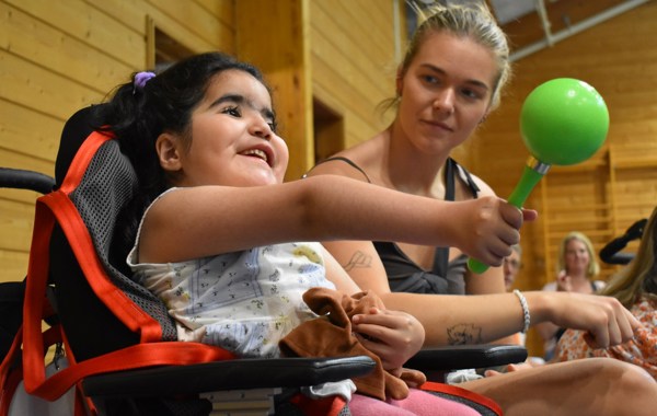 Jente med svart hår sitter i rullestol og holder en marakas, instrument, i hånda. En ung kvinne sitter ved siden av. 