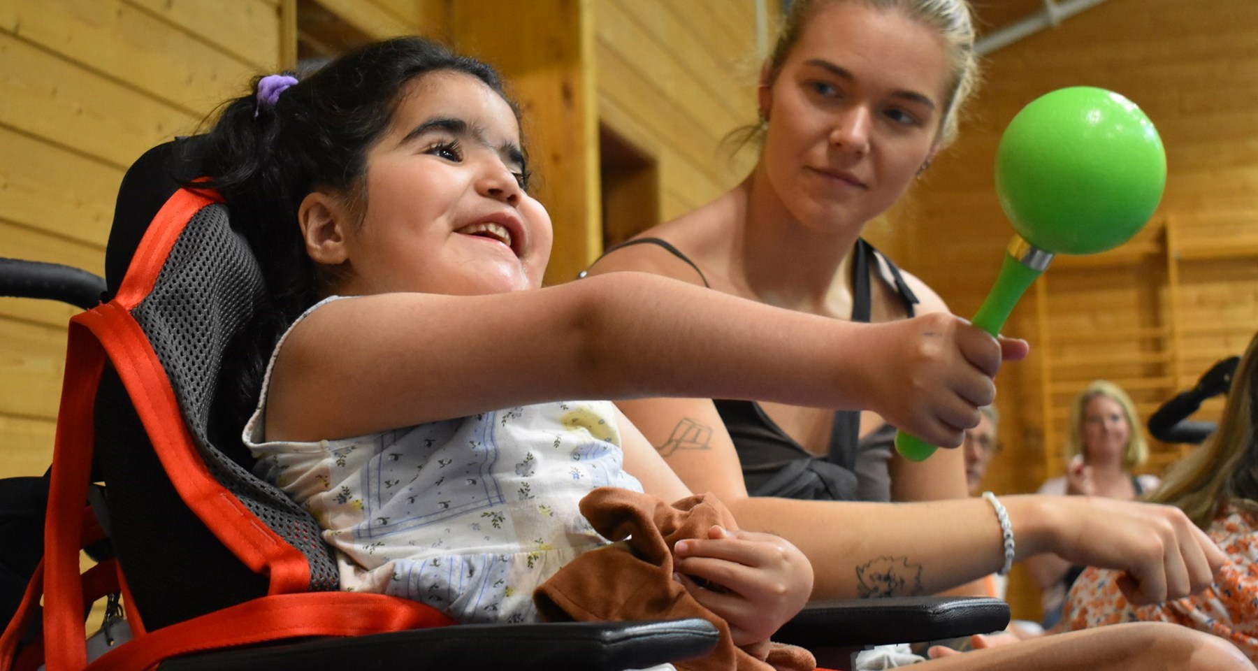 Jente med svart hår sitter i rullestol og holder en marakas, instrument, i hånda. En ung kvinne sitter ved siden av. 