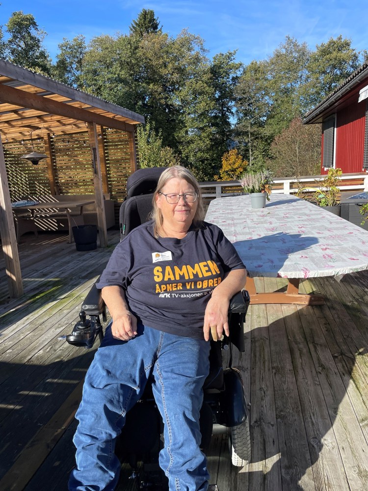 Kvinne sitter i rullestol ute på en terrasse. Hun har på seg TV-aksjons-trøye med skriften Sammen åpner vi dører. 