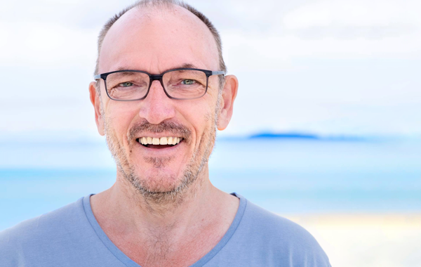 Mann med skjegg og briller, han smiler til kamera. Bakgrunnen er blå.