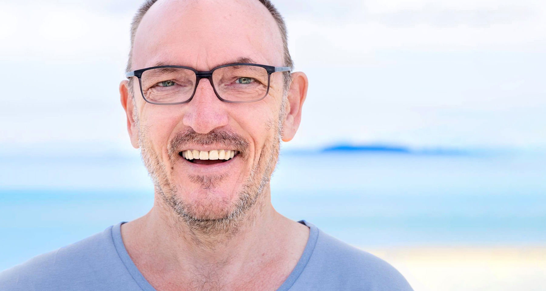 Mann med skjegg og briller, han smiler til kamera. Bakgrunnen er blå.