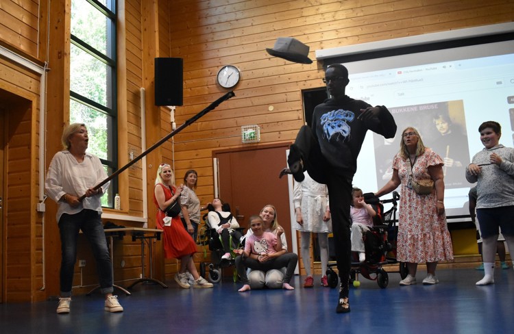 En gutt danser halling og tar hallingkast, han sparken hatten av en stokk. Barn og voksne står rundt i gymsalen, noen barn sitter i rullestol. 