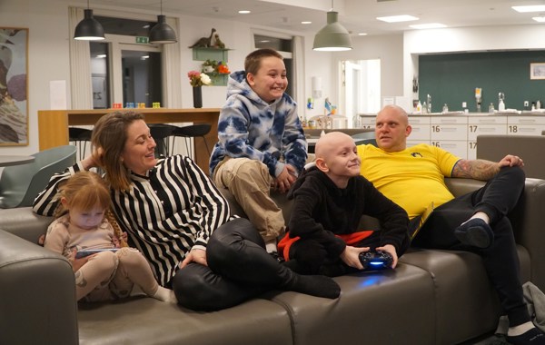 Familie med tre barn sitter i en sofa. To av dem spiller på konsoller. 