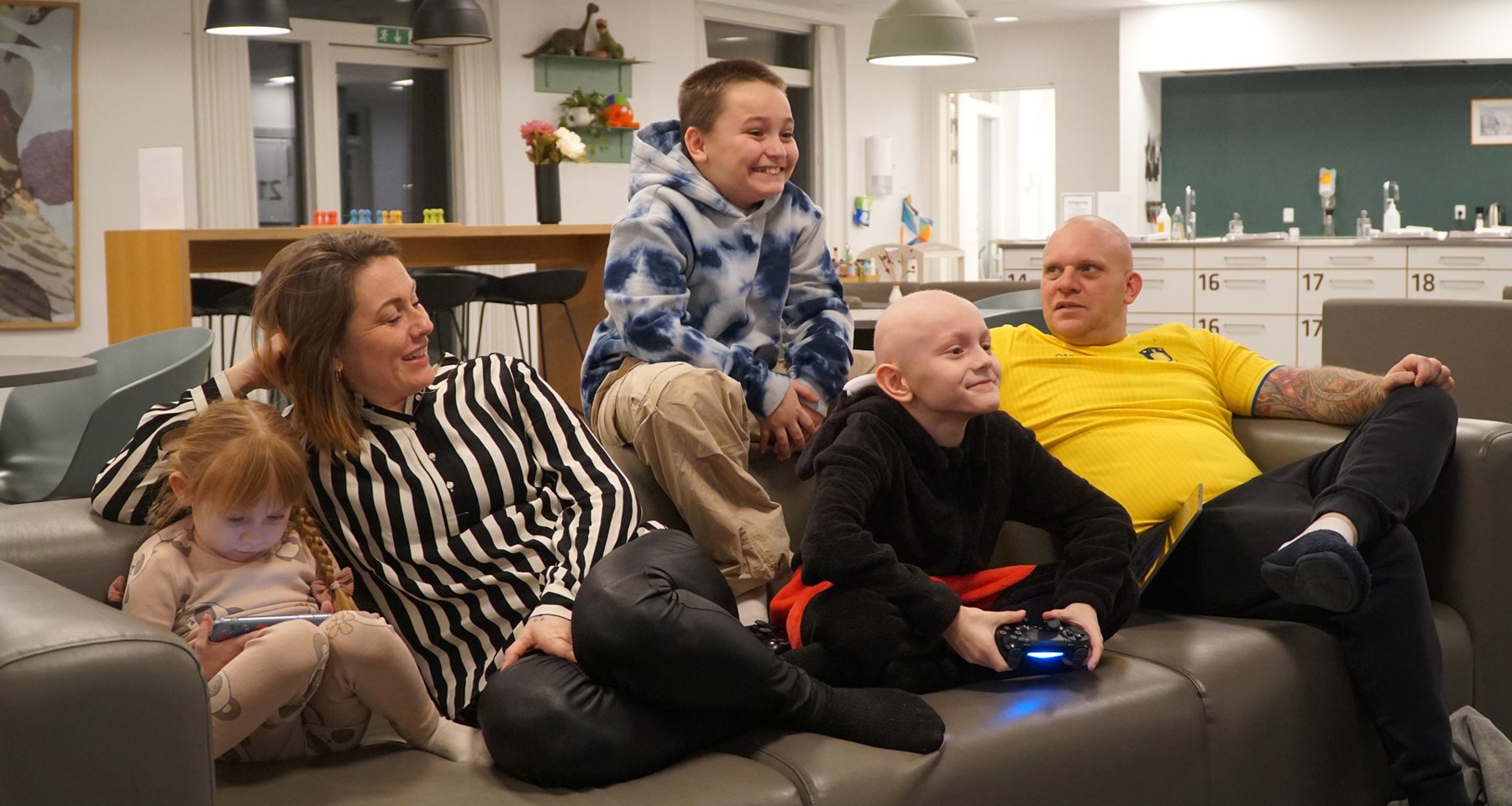 Familie med tre barn sitter i en sofa. To av dem spiller på konsoller. 