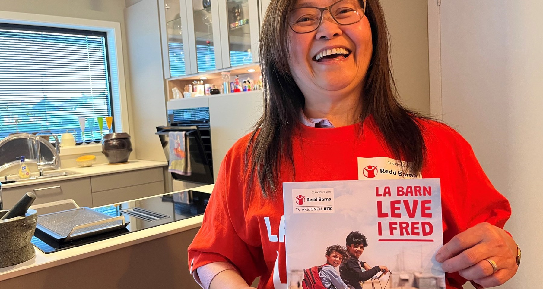 En kvinne med briller har på seg t-skjorte, og viser plakat, fra årets TV-aksjon.