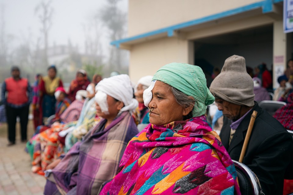 Eldre kvinner med fargerike klær sitter med lapp foran øyet utenfor en klinikk i Nepal. 