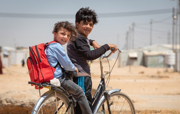 To barn sitter på en sykkel. Det ene barnet har på seg en rød sekk. 