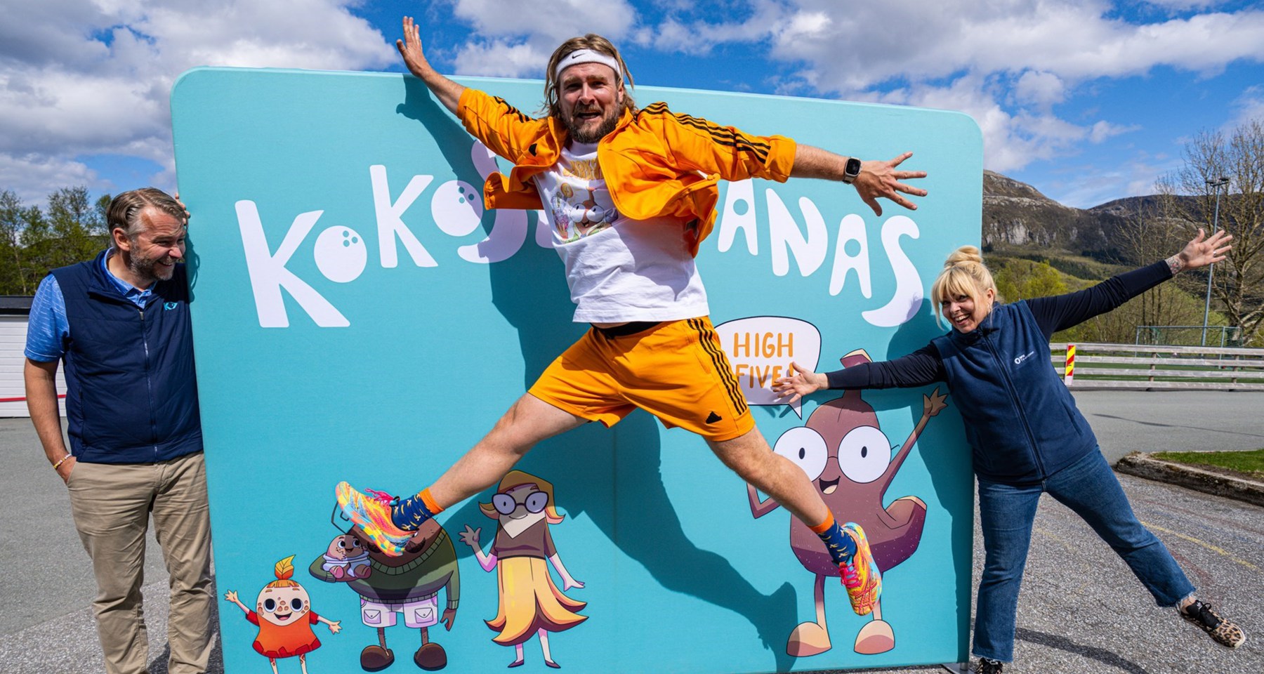 En mann hopper foran et skilt, holdt oppe av en kvinne og en mann.