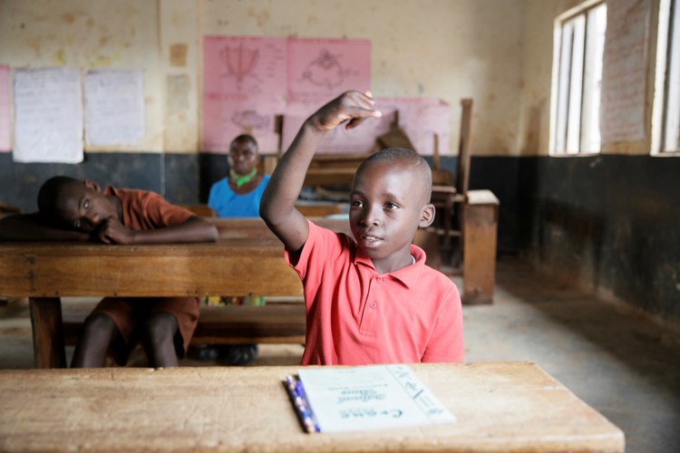 Gutt i rød trøye sitter i klasserom i Uganda. Han løfter opp armen og lærer tegnspråk. 