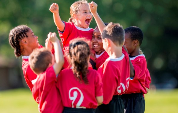 En gruppe barn i fotballdrakter feirer og strekker hendene i været.