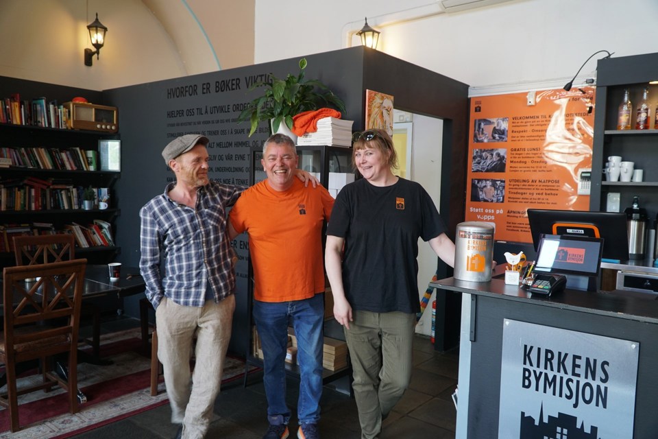 To menn og en kvinne står på Kirkens Bymisjons lokaler. To av dem i bymisjonens T-skjorter.