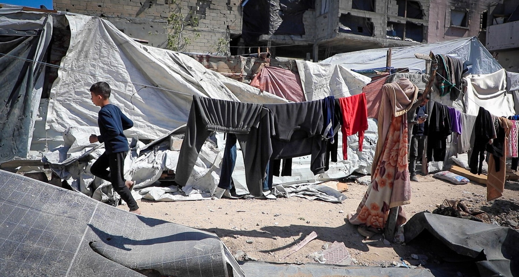 En ung gutt løper på en grusvei omringet av telt og klær på tørkesnorer. I bakgrunnen er det et murhus med store skader. 