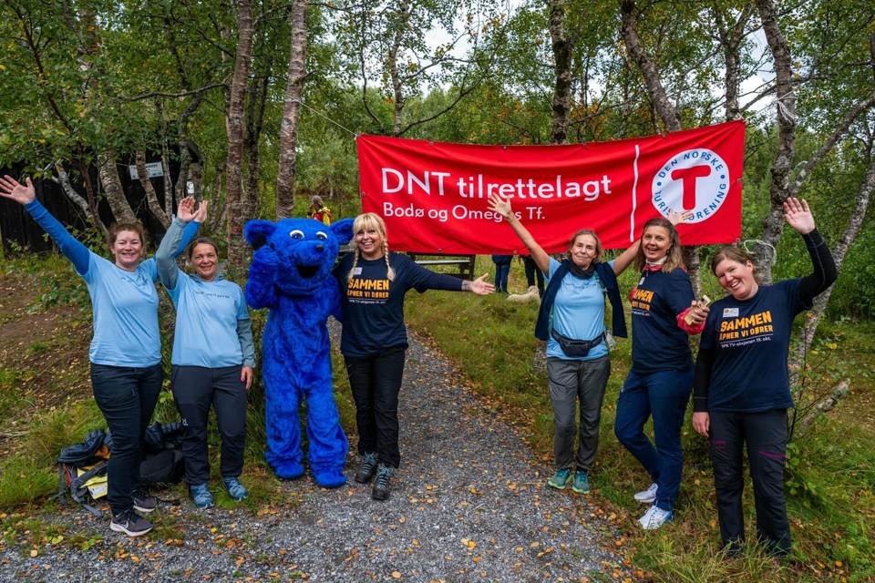 En gruppe mennesker er på tur i skogen. Et stort rødt banner med teksten DNT tilrettelagt henger mellom to trær.