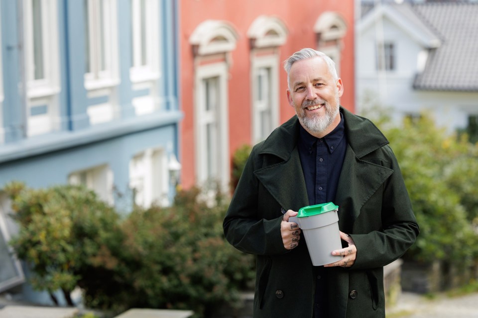 En mann med skjegg og svart frakk står med bøsse utenfor et hus. 