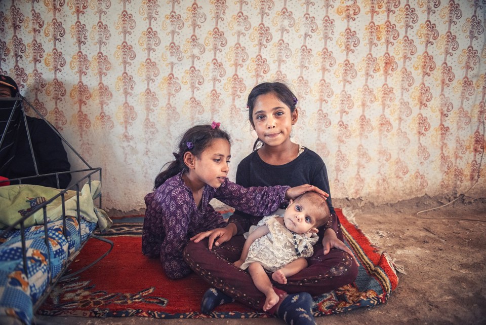 Tre barn sitter på bakken. Det er ikke gulv der, de sitter på et teppe. Den minste ungen er underernært.