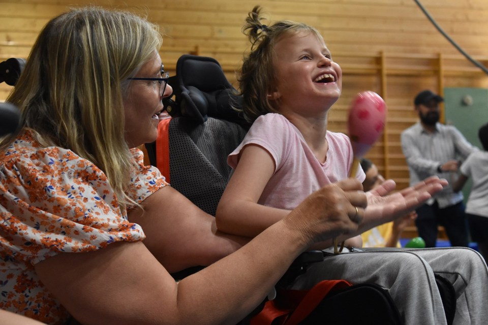 Jente med rosa trøye sitter i rullestol med blond kvinne ved siden av. Jenta smiler og ser glad ut. Kvinnen spiller instrumentet marakas.