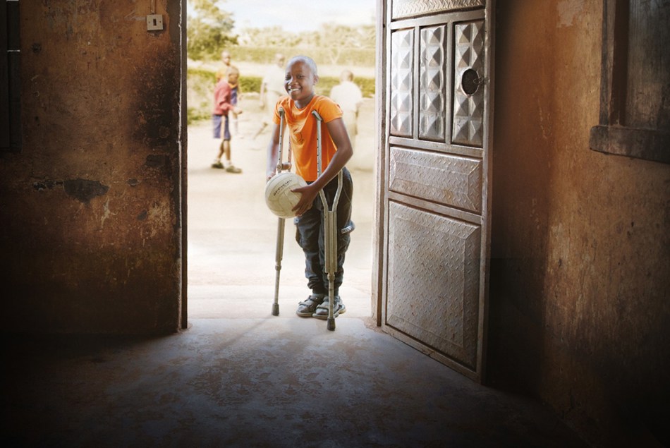 Mørkhudet gutt står i døråpning med krykker og en ball i hendene