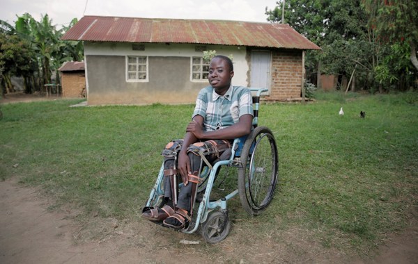 En ung, smilende gutt sitter i rullestol på gressplen med hus i bakgrunnen i Uganda. 