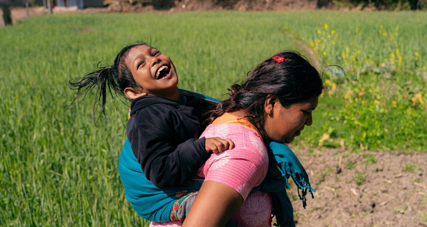 Jente sitter på ryggen til moren sin i en grønn åker i Nepal. Jenta smiler og ler. 