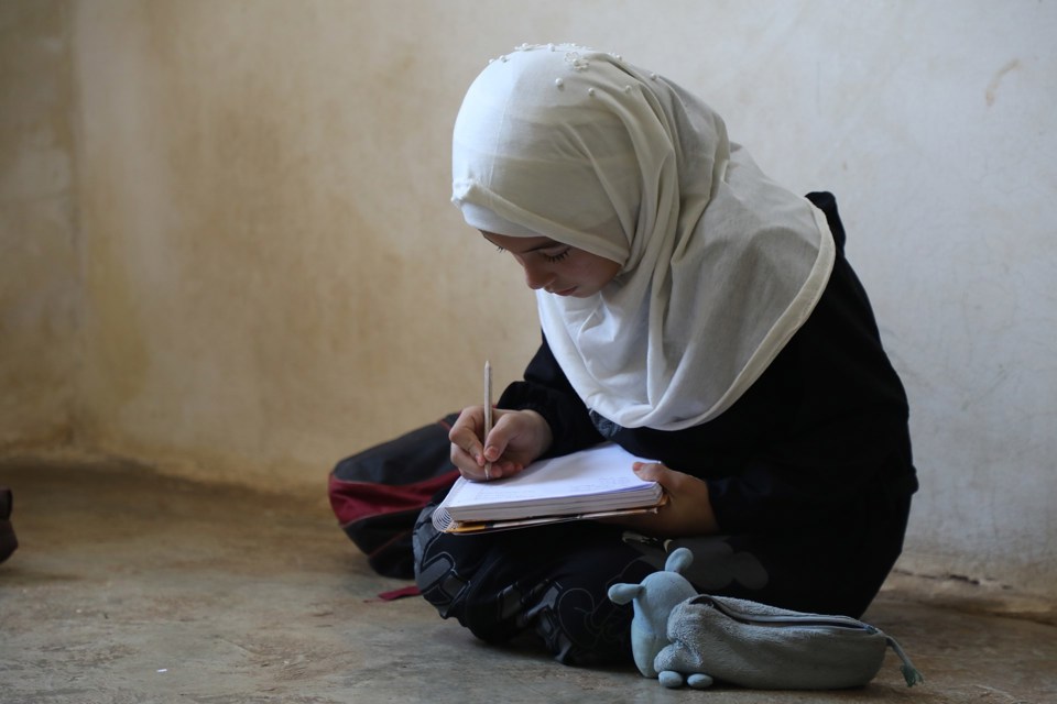 Jente gjør lekser sittende på bakken. Hun noterer i en skrivebok, og har et pennal ved siden av seg.