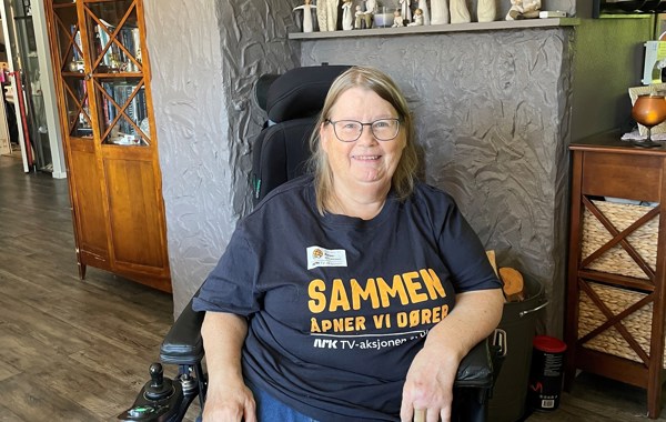 Kvinne sitter i rullestol inne i en stue. Hun har på seg TV-aksjons-trøye med skriften Sammen åpner vi dører. 