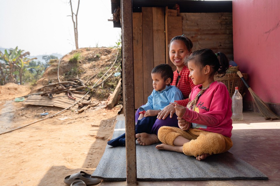 En kvinne og to barn sitter på en matte utenfor et hus. 