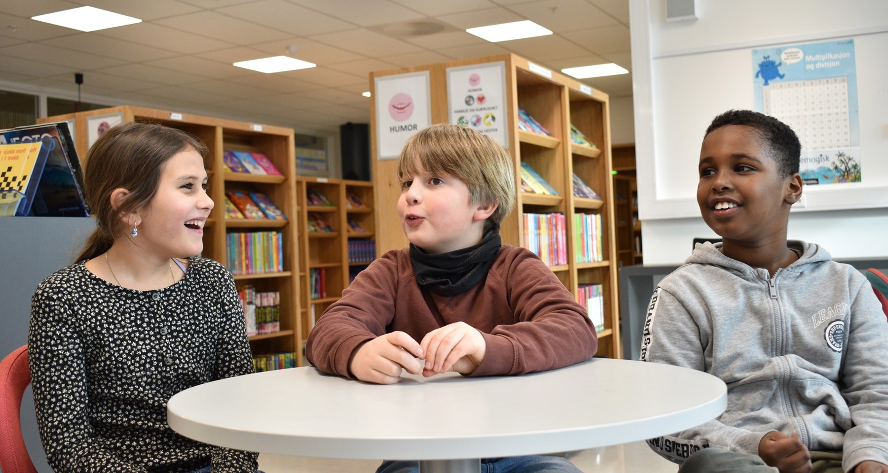 Tre unge elever sitter på et bibliotek, og smiler og snakker. 