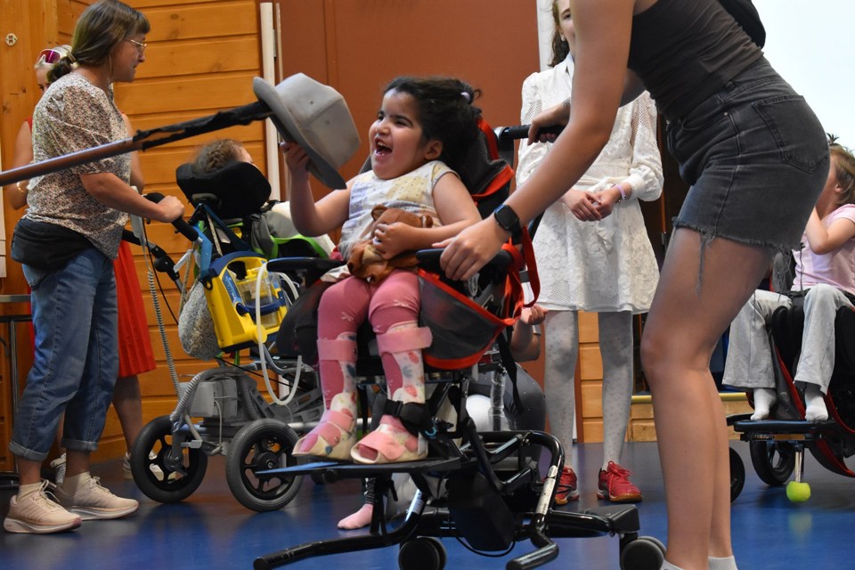 Liten jente i rullestol tar hatten av en stokk og smiler bredt. En voksen hjelper henne med hallingkast. 