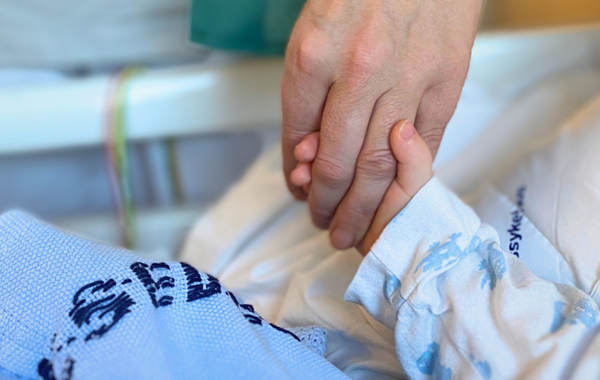 En voksen hånd som holder hånda til et barn som ligger i en sykeseng.