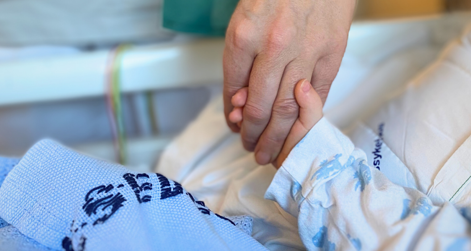 En voksen hånd som holder hånda til et barn som ligger i en sykeseng.