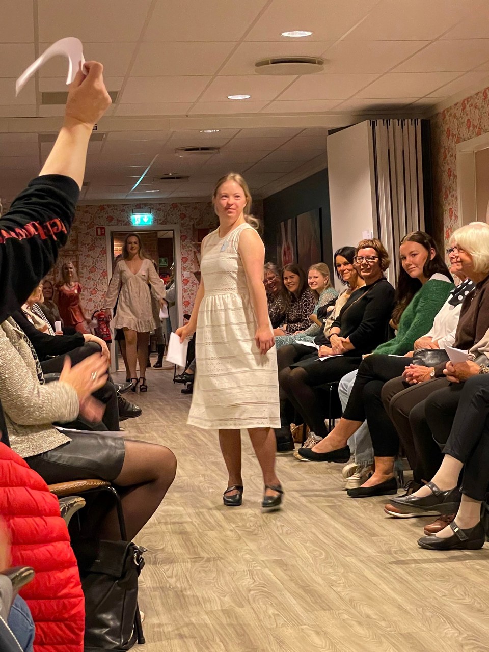 Kjoleauksjon for TV-aksjonen. To kvinner iført kjoler går i en midtgang, mens en rekke kvinner sitter på stoler på sidene.