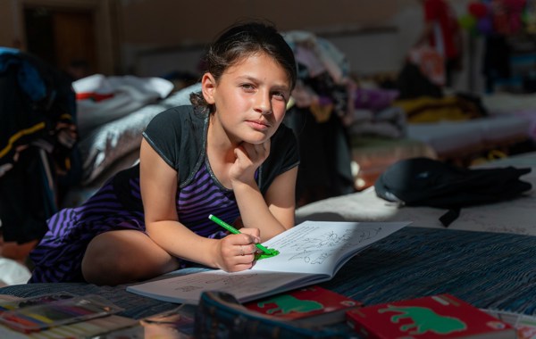 Ung jente sitter på gulvet og ser i kameraet. Hun har en skrivebok foran seg, og fargelegger med en grønn tusj.