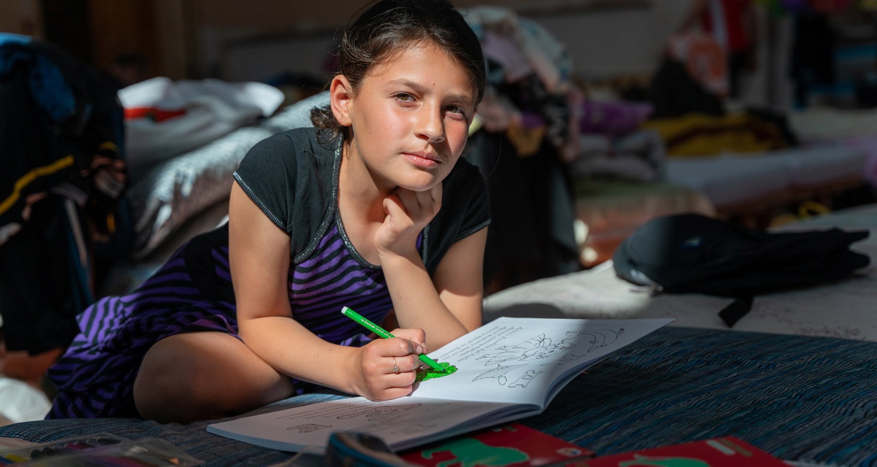 Ung jente sitter på gulvet og ser i kameraet. Hun har en skrivebok foran seg, og fargelegger med en grønn tusj.