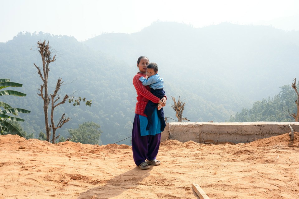 En kvinne står på bakke av sand med en liten gutt i armene. Bak henne er en skogkledd dal. 