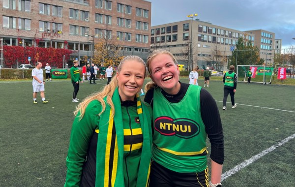 To jenter står på en fotballbane med grønne drakter og smiler. 