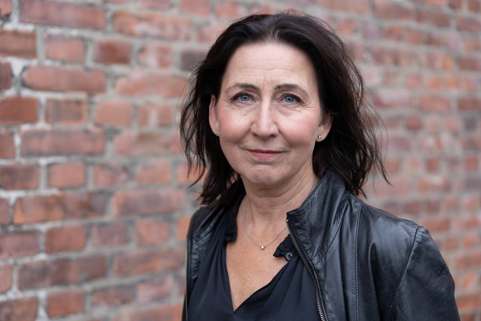 En kvinne med svart hår og svart skinnjakke, foran en murvegg.