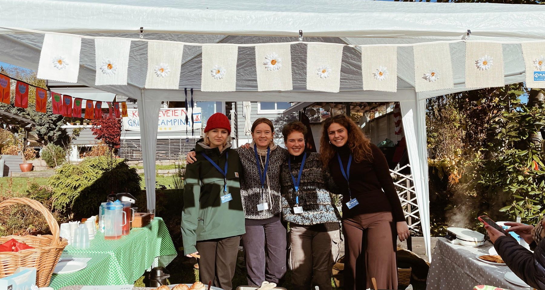 Fire jenter som står i et telt, med kaker og vafler på bord foran dem. De har kakesalg for TV-aksjonen. Det er pent gjort med duker og pynt.