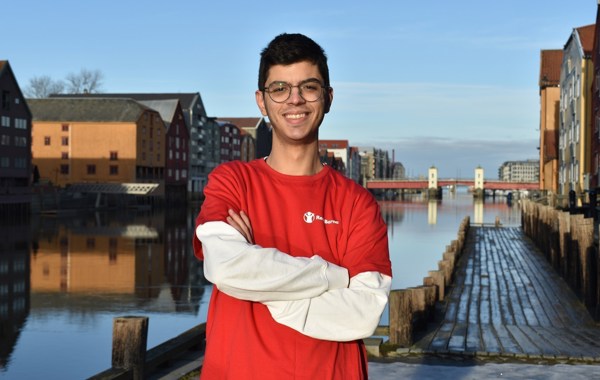 En gutt står på en brygge med vannet bak seg. Han har armene i kors, briller og smiler til kamera. Han har på seg en Redd Barna-tskjorte.