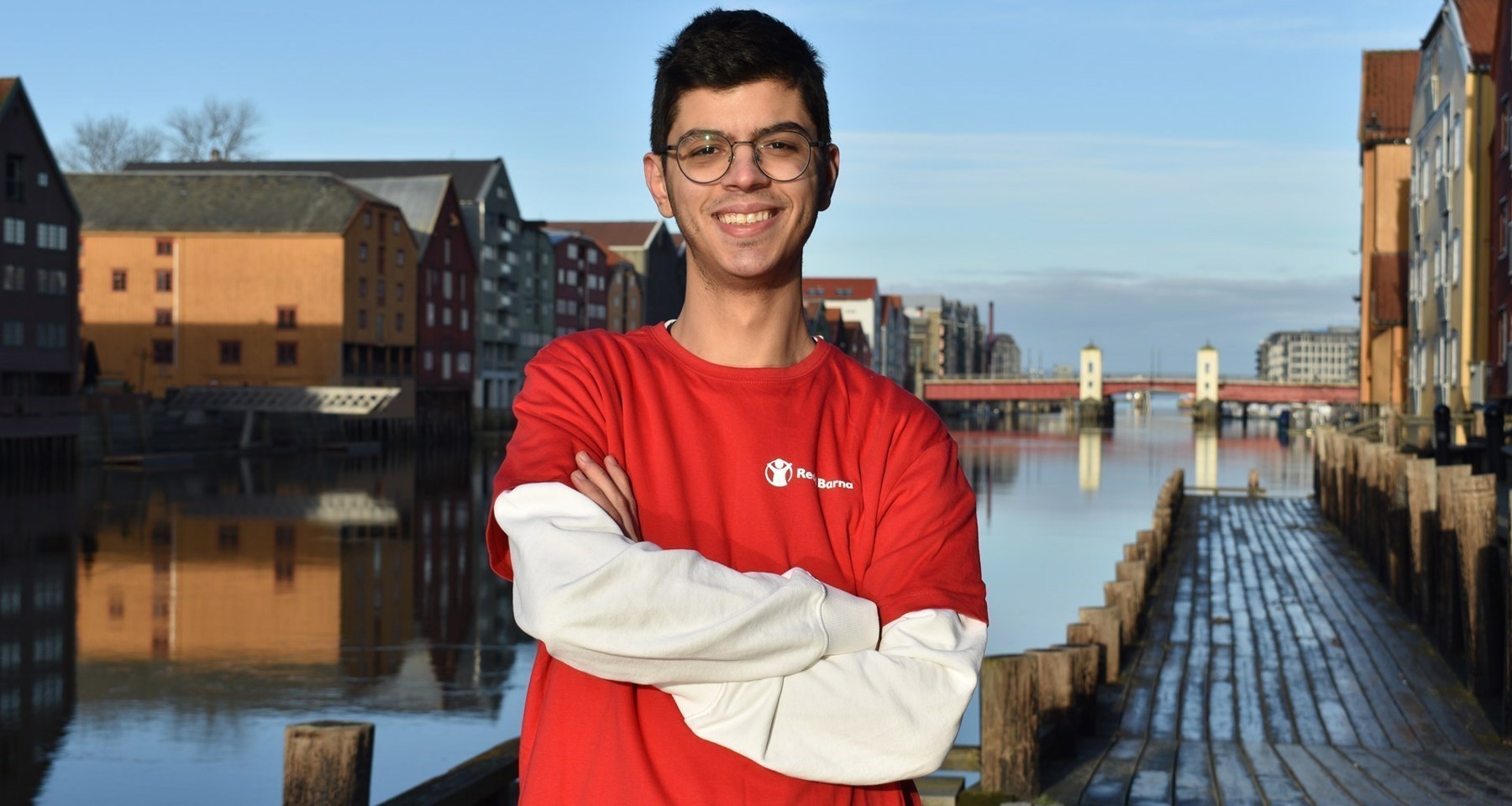En gutt står på en brygge med vannet bak seg. Han har armene i kors, briller og smiler til kamera. Han har på seg en Redd Barna-tskjorte.