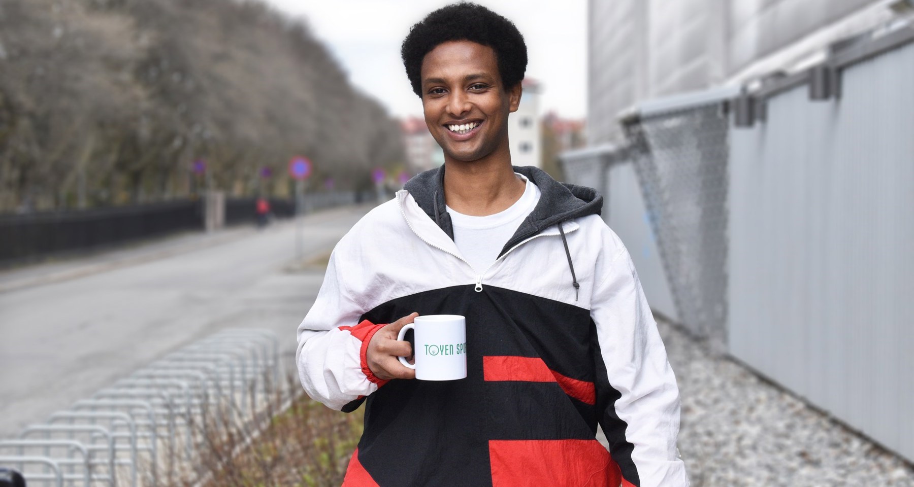En ung mann står foran et grått bygg, og smiler til kamera. Han har en kopp hvor det står "Tøyen sportsklubb" i hånda.