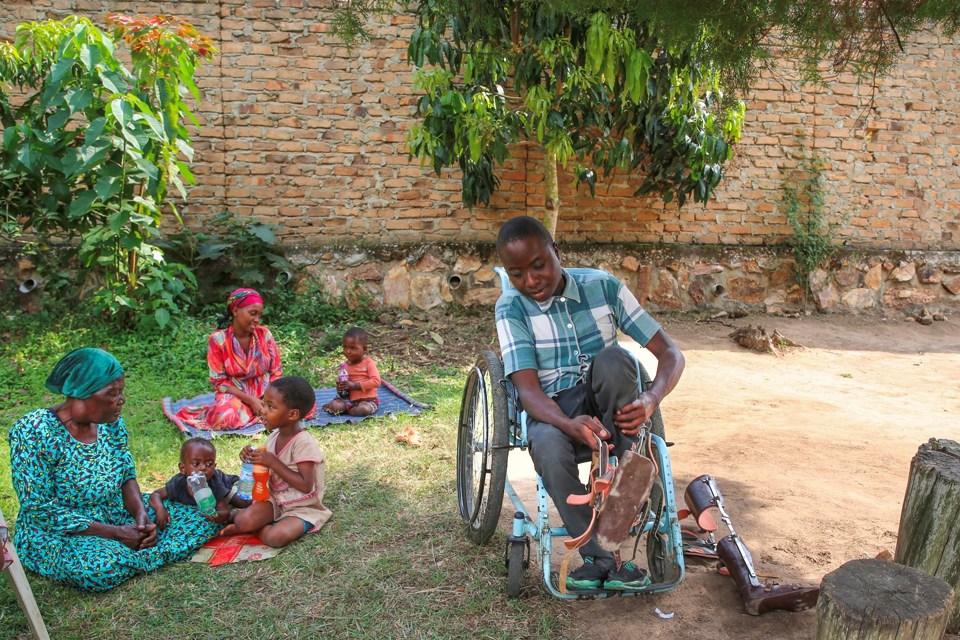 Gutt sitter i rullestol og tar på skinner på beina. Ved siden av sitter to kvinner og tre barn på plenen. 
