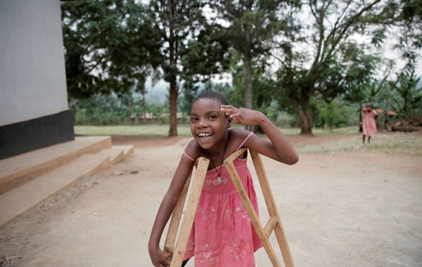 En smilende jente med rosa kjole står ute med krykker og løftet hånd. 