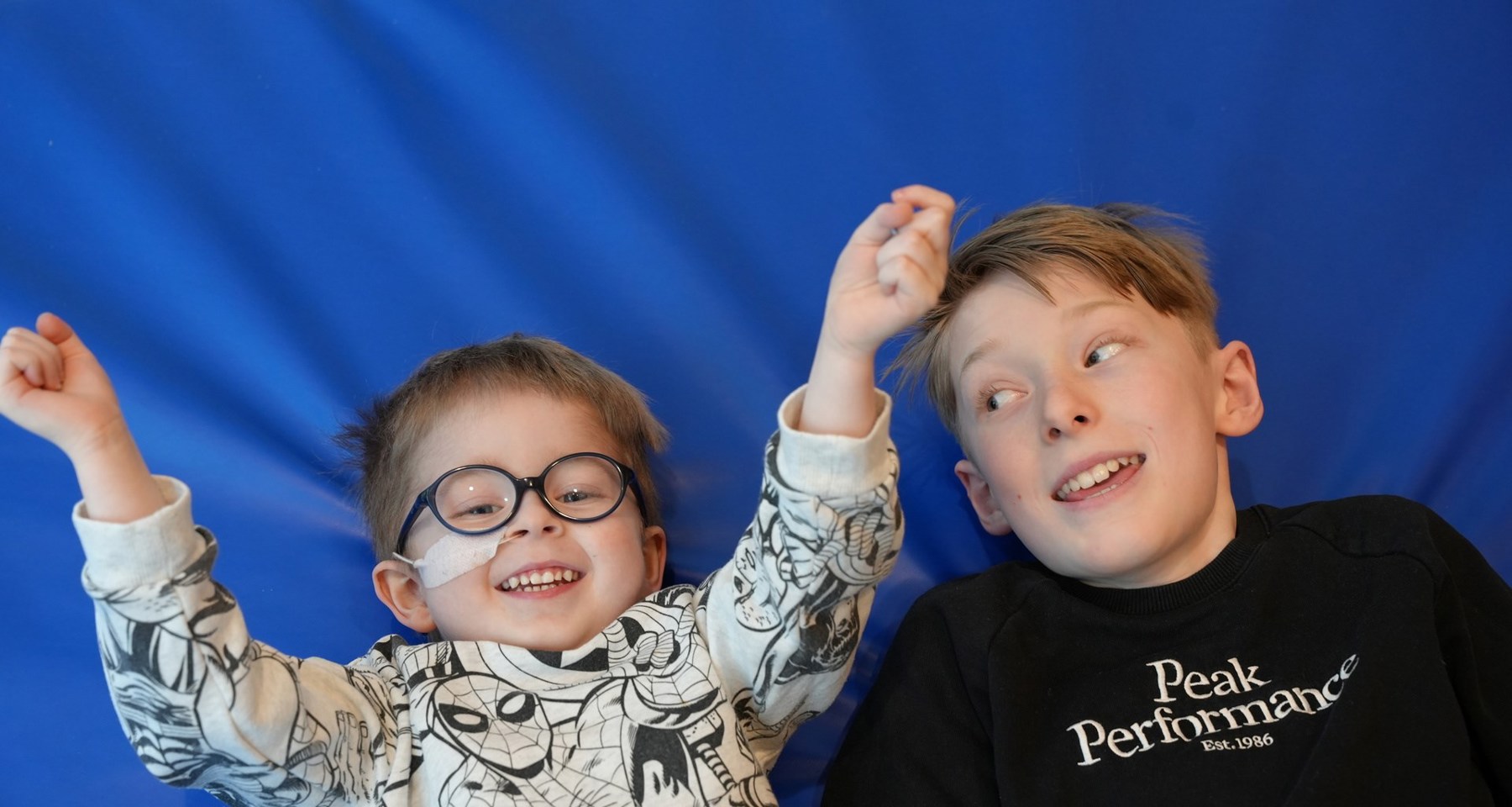 To unge gutter ligger ved siden av hverandre på blått underlag. En av dem har briller og nesesonde.